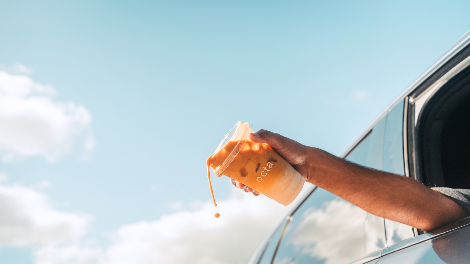 An iced Octa coffee held out of a car window against open sky.